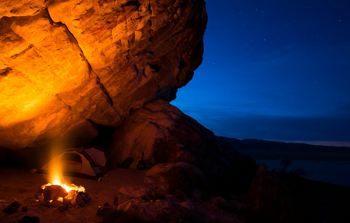 A glowing campfire sits beside a tent beneath a rock overhang.